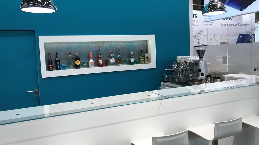 Weiße Messebar vor türkisfarbener Standwand, mit Siebträgermaschine und beleuchtetem Flaschenregal in der Wandnische