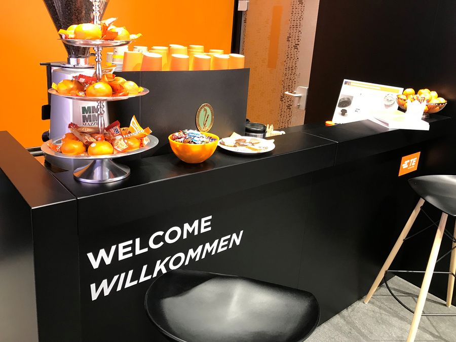 Schwarze Messetheke mit oranger Front und dem Schriftzug „Welcome Willkommen“, darauf Obst-Etageren und Gläser mit Orangensaft