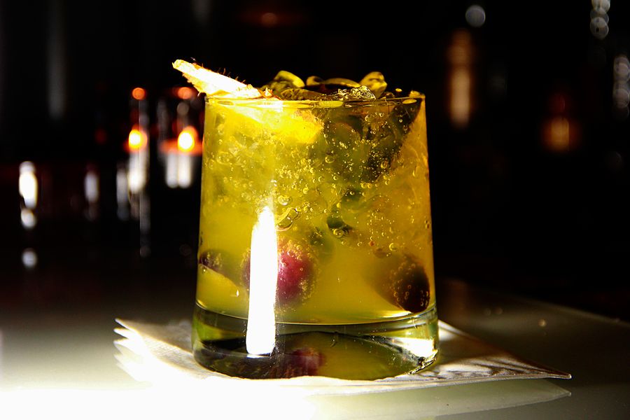 Gelbgrüner Cocktail im Tumbler mit Crushed Ice, Beeren und Kräutern vor dunkler Barkulisse