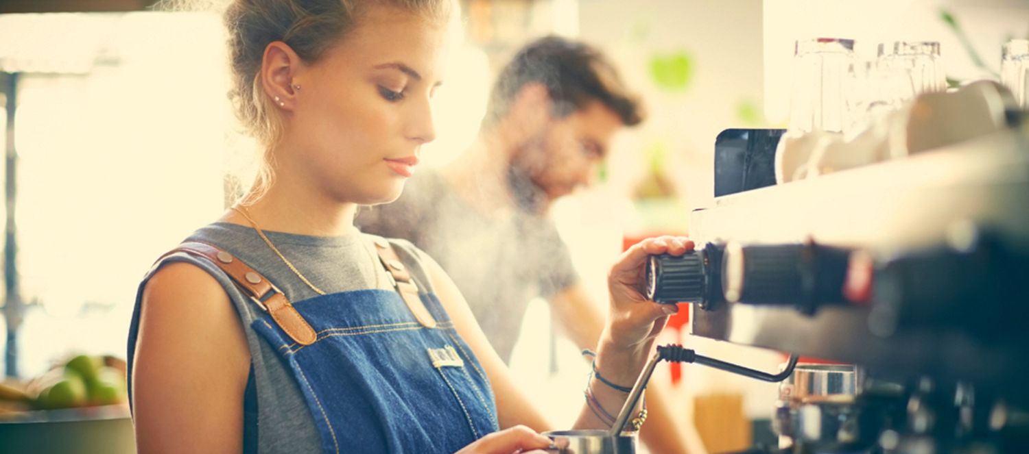 Barista stellt die Mühle an einer Siebträgermaschine ein, im Hintergrund dampfende Milch
