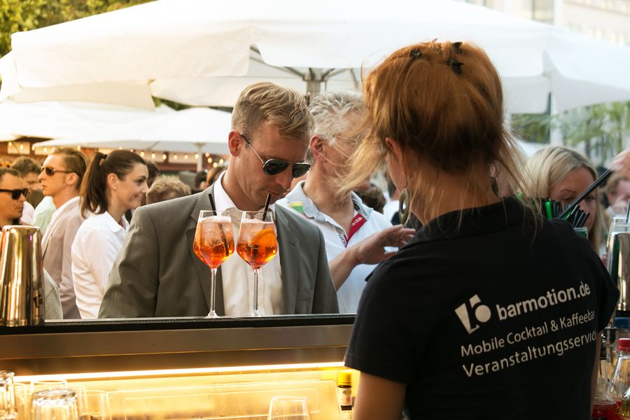 Servicekraft im barmotion-Shirt reicht zwei Drinks über die Theke einer Open-Air-Bar