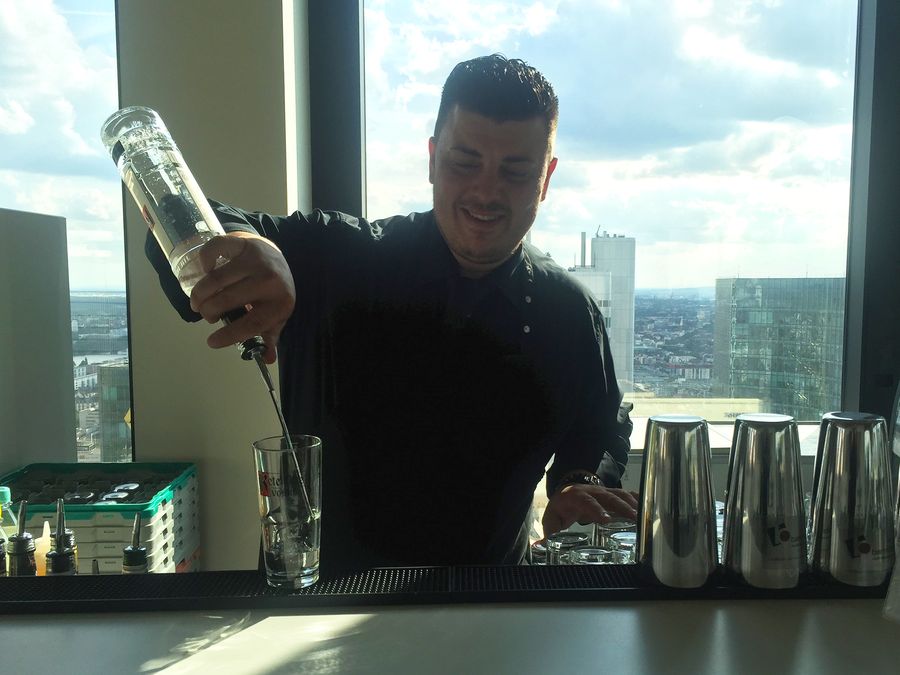 Barkeeper von barmotion gießt vor einer Fensterfront mit Skyline einen Drink ein
