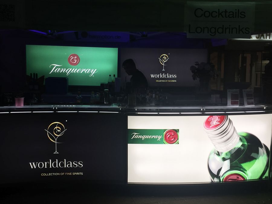 Beleuchtete Bartheke mit Rückwand voller Spirituosenflaschen und Schildern für Cocktails und Longdrinks